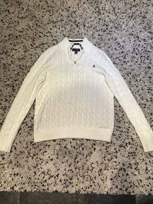 Vit kabelstickad tröja Polo Ralph Lauren - Vit kabelstickad tröja från Polo Ralph Lauren med halv dragkedja och ribbad krage med svarta ränder. Tröjan har lång ärm och klassisk broderad logga på bröstet. Perfekt för en stilren och sportig look.