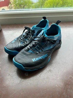 Svarta och blåa inomhusskor från Kempa - Säljer ett par sportiga inomhusskor från Kempa i svart och blått. Skorna har snörning, låg profil och är tillverkade i ett lätt och ventilerande material. Perfekta för handboll eller annan inomhussport. Snygg design med Kempa-logga på sidan. Säljer på grund av att dem aldrig är använda, alltså superfräscha. Storlek 39.