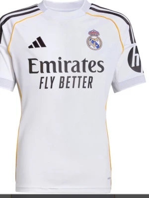 Real Madrid matchtröja Vini Jr. 7  vilken storlek som helst  - Vit Real Madrid matchtröja från Adidas med svarta detaljer, klubbmärke och sponsortryck på bröstet. Nummer 7 och VINI JR. på ryggen. Korta ärmar, gula inslag längs sidorna och axlarna. Perfekt för fotbollsfans som vill visa sitt stöd.