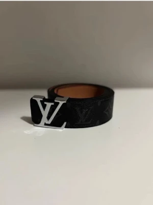 Svart bälte med monogram - Stilrent svart bälte från med klassiskt silverfärgat. Bältet har en elegant design och är tillverkat i skinn med diskret mönster. Perfekt accessoar för att lyfta din outfit.