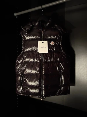 Svart dunväst från Moncler - Snygg svart dunväst från Moncler med glansig finish och klassisk logotyp på bröstet. NFC tillgängligt på loggan. Västen har dragkedja framtill, hög krage och två fickor med dragkedja. Perfekt för lager-på-lager och streetstyle. Materialet är dunfyllt och yttermaterialet är blankt.