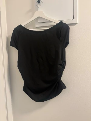 Svart topp med spets och rosett - Snygg svart topp med korta ärmar, djup rygg med spetsdetaljer och en stor rosett bak. Toppen har en feminin och trendig look, perfekt att styla med jeans eller kjol. Materialet känns mjukt och följsamt.