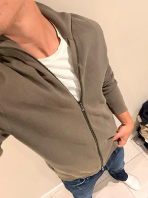 Olivgrön hoodie med dragkedja - Snygg olivgrön hoodie med dragkedja framtill och huva. Tillverkad i mjukt bomullsmaterial, perfekt för en avslappnad stil. Passar bra till jeans eller joggers.