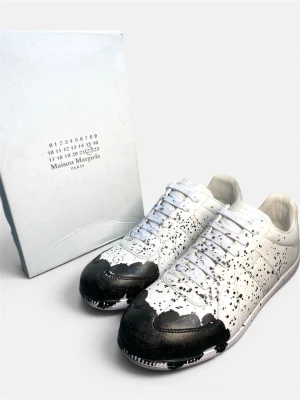 Maison Margiela vita sneakers med svart färgstänk - Maison Margiela sneakers i vitt läder med unikt svart färgstänksmönster och svarta detaljer på tån. Klassisk låg modell med vita skosnören och vit sula. Perfekt för dig som gillar statement-plagg och vill sticka ut med din stil.