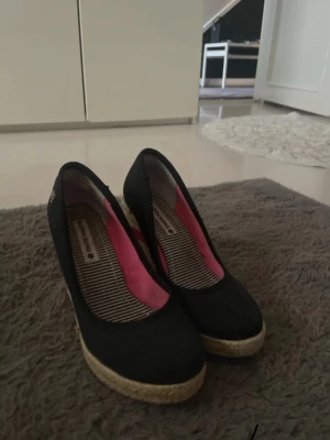 Svarta kilklackade pumps från Bianco - Snygga svarta pumps från Björn borg med kilklack i flätad jute och rosa detaljer på insidan. Skorna har rund tå och randigt innerfoder. Perfekta för dig som gillar en mix av klassiskt och trendigt.