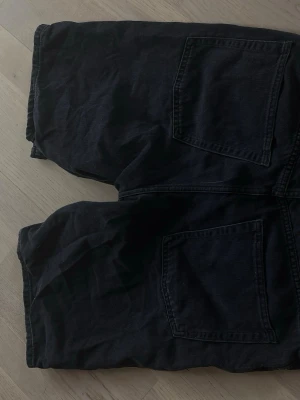 Svarta denimshorts med stora fickor - Säljer ett par svarta denimshorts med stora bakfickor och klassisk femficksdesign. Shortsen har en rak passform och stängs med dragkedja och knapp framtill. Perfekta för en avslappnad stil under sommaren.