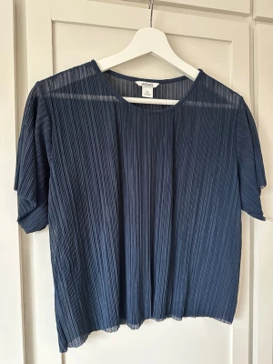 Mörkblå plisserad topp från Monki XS - Snygg mörkblå plisserad blus från Monki med korta ärmar och rund halsringning. Tillverkad i ett lätt, transparent material som ger en luftig känsla. Perfekt att styla med jeans eller kjol för en trendig look.