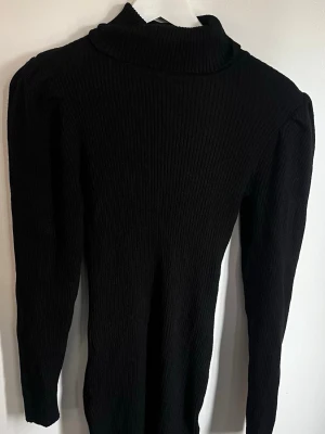 Svart poloklänning ner till knäna - Säljer en svart poloklänning med lång ärm och puffade axlar. Klänningen har en tight passform och hög krage som ger en stilren look. 