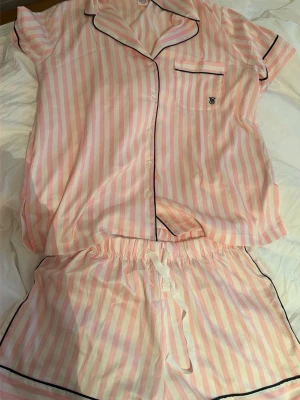 Randig rosa pyjamas från Victoria's Secret - Säljer en kortärmad pyjamas från Victoria's Secret med rosa och vita ränder. Setet består av en skjorta med knappar, svart passpoal och bröstficka samt matchande shorts med dragsko i midjan. Perfekt för mysiga kvällar och morgnar.