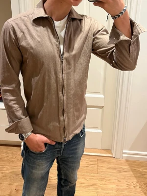 Brun skjorta med dragkedja John Henric - Säljer en stilren brun skjorta/overshirt från John Henric med klassisk krage och hel dragkedja framtill (uppifrån och nedifrån). Tillverkad i ett lätt tyg som passar perfekt nu till sommaren☀️Inga defekter. Storlek S. Skriv vid minsta fundering!