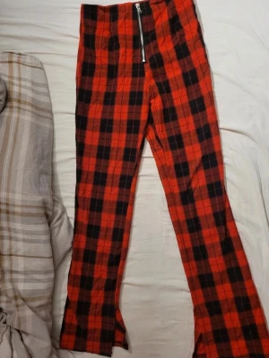 Rödrutiga leggings med dragkedja - Snygga leggings med rött och svart rutmönster och en synlig dragkedja framtill. Hög midja och utsvängda ben ger en trendig look. Perfekta för dig som vill sticka ut med din stil.