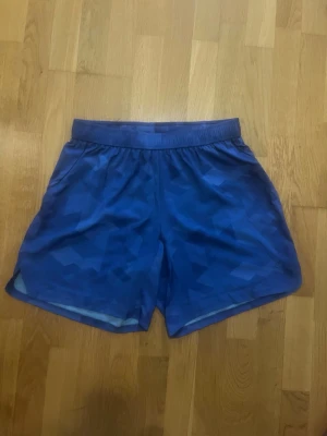 Blå träningsshorts med mönster - Snygga blå träningsshorts med geometriskt mönster och elastisk midja. Shortsen har en liten dragkedjeficka bak och är tillverkade i ett lätt och svalt syntetmaterial, perfekta för aktiva dagar.