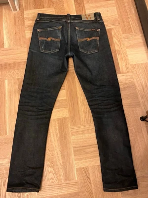 True Selvedge jeans från Nudie Jeans - Säljer ett par selvedge jeans från Nudie Jeans med klassisk femficksdesign och orange kontrastsömmar. Jeansen har raka ben och Nudie-loggan broderad på bakfickorna. Tillverkade i kraftig denim, perfekta för en stilren look.