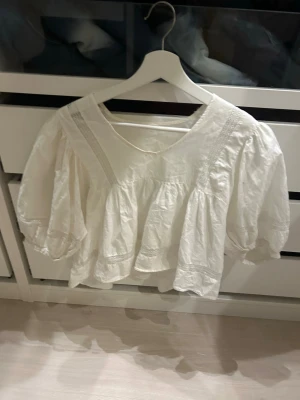 Vit volangblus med puffärm - Superfin vit blus med volanger och breda puffärmar. Blusen har v-ringning och dekorativa spetsdetaljer längs axlar och framstycke. Luftig och lös passform, perfekt till jeans eller kjol. Materialet känns som bomull.