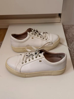 Vita sneakers från K.Cobler - Snygga vita sneakers från k.Cobler med klassisk låg profil och beige sula. Skorna har guldfärgade öljetter, diskret logga på sidan och är tillverkade i läder med textildetaljer på insidan. Perfekta för en clean och stilren look.
