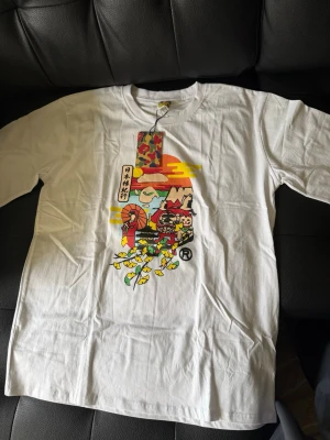 Vit Bape t-shirt med japanskt tryck - Vit t-shirt från Bape med färgglatt japanskt tryck framtill. Motivet har inslag av kamouflage och tecknade figurer. Klassisk passform och rund halsringning. Perfekt för dig som gillar streetwear och unika prints.