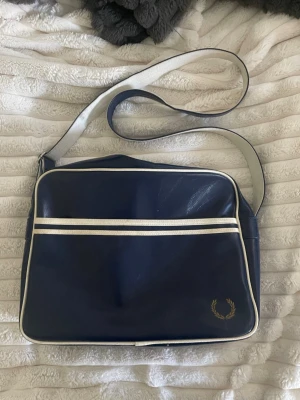 Mörkblå axelväska från Fred Perry - Snygg marinblå äkta axelväska från Fred Perry med vita detaljer och justerbar axelrem. Väskan har klassisk retrostil och vita detaljer längst dragkedjan. Den är knappt använd av mig men är lite smutsig på remmen, syns i bilden, men går säkert att tvätta bort.  Skriv gärna för fler frågor 💕Pris kan diskuteras  Mått: 38 x 11 x 26 (cm)