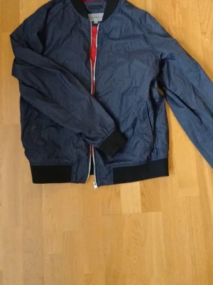 Blå bomberjacka från Calvin Klein Jeans - Snygg blå bomberjacka från Calvin Klein Jeans med svart ribbad krage, muddar och nederkant. Jackan har röd insida och dragkedja framtill. Perfekt för dig som vill ha en stilren och klassisk look. Storlek large men sitter som en small/medium!