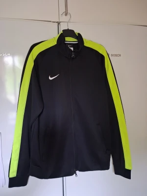 Nike svart träningsjacka med neonränder - Svart träningsjacka från Nike med dragkedja och neonfärgade ränder längs ärmarna. Jackan har ståkrage, två fickor framtill och klassisk vit Nike-logga på bröstet. Perfekt för träning eller sportiga outfits. Storlek L