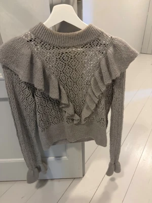 Beige stickad tröja med volanger - Trendig beige stickad tröja med volangdetaljer och hålmönster. Tröjan har lång ärm, ribbade muddar och en högre halsringning. Perfekt för dig som gillar en romantisk och unik stil.