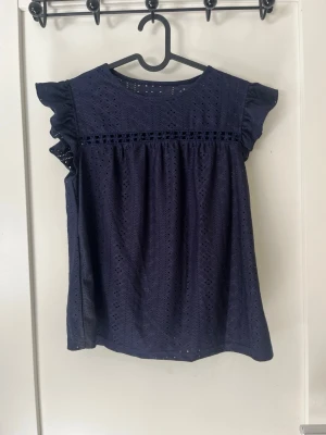 Mörkblå spetsblus med volangärm - Supersöt mörkblå blus med broderat spetsmönster och volangdetaljer vid ärmarna. Blusen har rund halsringning och en dekorativ bård framtill. 