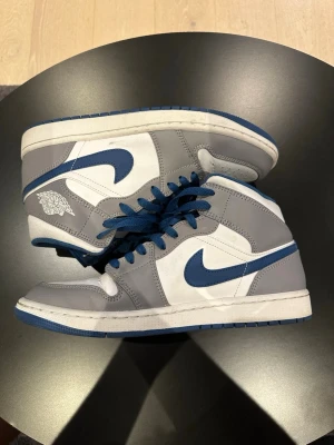 Air Jordan 1 Mid grå/blå sneakers - Snygga Air Jordan 1 Mid sneakers i grått och vitt med blå detaljer och blå swoosh. Skorna har högt skaft, blå skosnören och klassisk Jordan-logga på sidan. Ovandelen är i läder och mesh för en clean och sportig look.