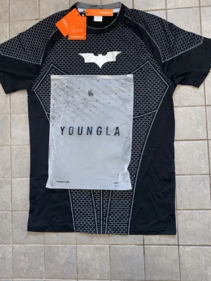 Batman-inspirerad t-shirt från YoungLA - Svart Batman-inspirerad t-shirt från YoungLA med vit batmanlogga på bröstet och hexagonmönster över hela framsidan. Tröjan har korta ärmar och är tillverkad i ett stretchigt, funktionsmaterial som passar perfekt för cosplay eller maskerad.