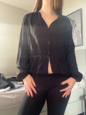 Svart blus med knappar och knytband - Snygg svart blus med knappar framtill och ett knytband i midjan som ger en feminin siluett. Blusen har v-ringning och långa ärmar med lätt puff vid axlarna. Perfekt att styla med jeans eller kjol för en trendig look.