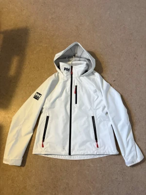 Vit vindjacka från Helly Hansen - Snygg vit vindjacka från Helly Hansen med huva och svarta dragkedjor. Ny pris 1500kr Tre praktiska fickor med dragkedja. Perfekt för blåsiga dagar och sportiga outfits, skyddar även vid regn. Inget är fel på skicket och inga slitage, nån/några småfläckar (bilden) som man inte tänker på