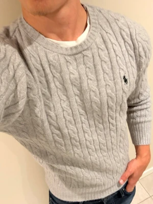 Ljusgrå kabelstickad tröja Polo Ralph Lauren - Snygg ljusgrå kabelstickad tröja från Polo Ralph Lauren med rund halsringning och klassisk broderad logga på bröstet. Tröjan har långa ärmar och ribbade muddar. Perfekt för lager-på-lager och enkel att matcha med jeans.