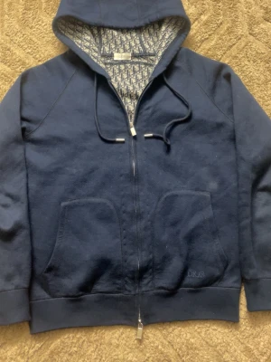 Mörkblå hoodie från Dio - Snygg mörkblå hoodie från Dio med dragkedja och två stora fickor framtill. Insidan av huvan har ett mönstrat Dio-tyg och loggan är diskret broderad på ena fickan. Mjuk bomullskvalitet och klassisk passform med dragsko i huvan.
