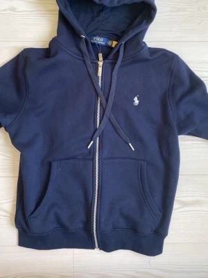 Mörkblå hoodie från Polo Ralph Lauren - Mörkblå hoodie med dragkedja från Polo Ralph Lauren. Klassisk design med huva, dragsnören och två fickor framtill. Ikoniska broderade loggan i vitt på bröstet. Perfekt för en avslappnad och stilren look.