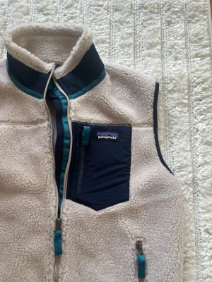 Patagonia beige fleeceväst - Säljer en beige fleeceväst från Patagonia med marinblå detaljer och dragkedjor. Västen har hög krage, en bröstficka med dragkedja och två sidofickor. Perfekt för lager-på-lager och outdoor-stil. Materialet är mjuk teddyfleece.