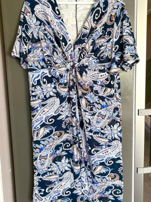 Klänning med knyt plussize - Snygg klänning med paisleymönster i blått, beige och brunt. Klänningen har v-ringning, korta ärmar och ett knyt framtill som ger en fin detalj. Skön, Stretchig kvalitet 