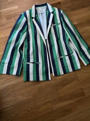 Randig kavaj från Pykettes - Snygg kavaj från Pykettes med breda vertikala ränder i grönt, blått och vitt. Klassisk krage, tre fickor framtill med helfodrad insida. Perfekt för dig som vill sticka ut med en färgglad och retroinspirerad look.