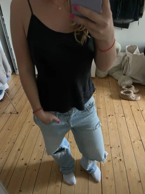 Svart silkestopp  - Snygg svart silkestopp med tunna axelband och avslappnad passform. Toppen är ärmlös och har en enkel, stilren design som passar perfekt till jeans eller kjol. Materialet känns mjukt och lätt mot huden. Justerbara band & små söta volanger/detaljer nedtill🙌Passar S-M