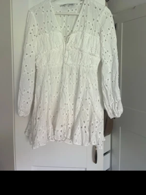 Vit kort klänning med broderade detaljer - Superfin vit kort klänning med broderade hålmönster och v-ringning. Klänningen har långa ärmar och volang nertill, samt smockad midja för en bekväm passform. Perfekt för varma dagar och somriga tillfällen.