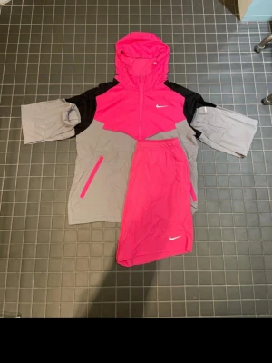 Nike Miller rosa set + shorts - Nike träningsset med jacka och shorts i stark rosa, grått och svart. Jackan har huva, dragkedja och fickor med rosa detaljer samt Nike-logga på bröstet. Shortsen är rosa med vit Nike-logga. Perfekt för löpning eller träning utomhus.