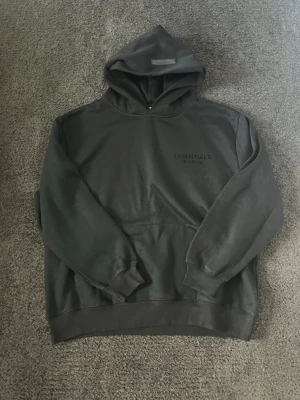 Svart Essentials Fear of God hoodie - Svart hoodie från Essentials Fear of God med diskret logga på bröstet. Klassisk passform med huva och magficka. Tillverkad i mjukt bomullsmaterial, perfekt för en avslappnad streetwear-look.