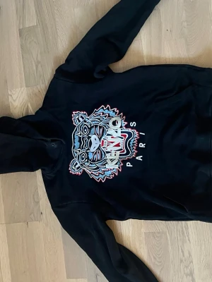kenzo hoodie - Svart hoodie från Kenzo med ikoniskt tigerhuvud broderat i flera färger på bröstet och texten 'KENZO PARIS'. Klassisk huva och mjukt material som känns skönt mot huden. Perfekt för dig som vill sticka ut med en streetwear-look.