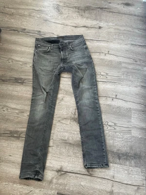 Grå slim fit jeans från Nudie st30/32 - Snygga grå slim fit jeans från Nudie med klassisk femficksdesign och svarta detaljer på bakfickorna. Storleken är 30/32. Jeansen har en något tvättad look och raka ben. Perfekta för en avslappnad och trendig stil. Dom har slittningar vid midjan och vi skrevet men inga hål och funkar som vanligt