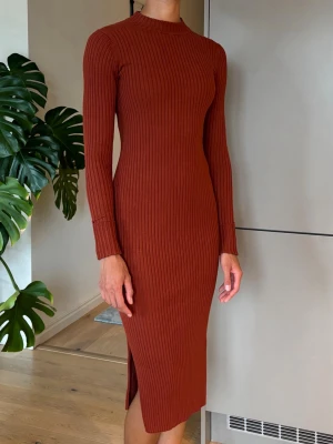 Rostbrun långärmad klänning från Monki  - Snygg ribbad klänning i rostbrun färg med lång ärm och slits på sidan. Klänningen har en kroppsnära passform och hög hals, plagget är stickat och känns mjukt mot huden. 