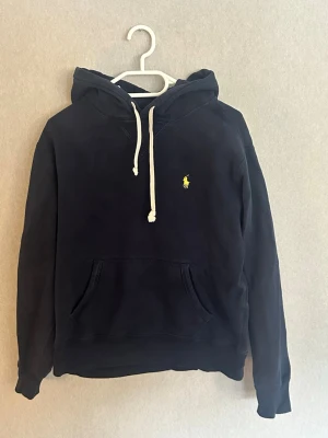 Mörkblå hoodie från Polo Ralph Lauren - Snygg mörkblå hoodie från Polo Ralph Lauren med klassisk gul logga broderad på bröstet. Hoodien har huva med vita dragsnören och en stor känguruficka framtill. Perfekt för en avslappnad och stilren look.