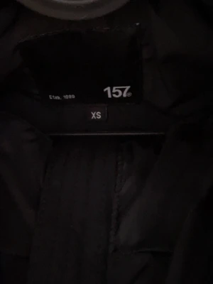 Svart lång dunjacka från 157 - Säljer en svart lång dunjacka från 157 i storlek XS. Jackan har huva och quiltad design som ger en snygg siluett. Perfekt för kalla vinterdagar och håller dig varm med sin vadderade insida. Enkel och stilren look som passar till det mesta.