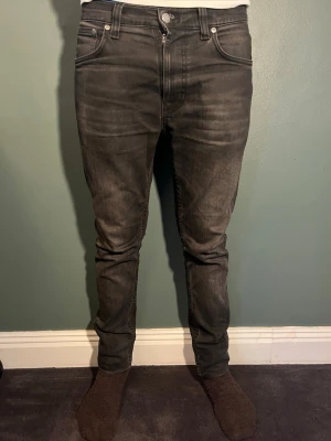 Svarta slim fit jeans från Nudie - Säljer ett par svarta slim fit jeans från Nudie Jeans med klassisk femficksmodell och snygga slitningar. Jeansen har en diskret logga på bakfickan och är tillverkade i mjuk denim med stretch för extra komfort. Nypris va 1600 kr