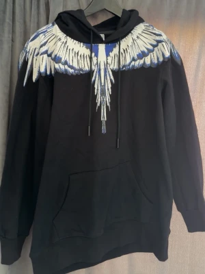 Svart hoodie med blå-vita vingar - Snygg svart hoodie med stort tryck av blå och vita vingar över axlar och huva, både fram och bak. Hoodien har en klassisk känguruficka och justerbar dragsko i huvan. Perfekt för dig som vill sticka ut med en unik design.