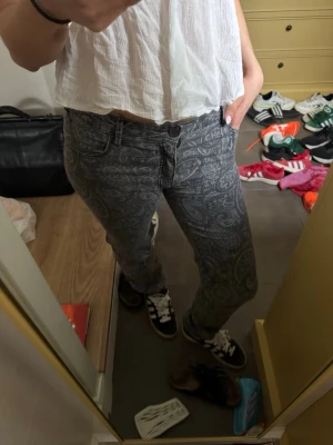 Grå paisleymönstrade jeans - Säljer ett par grå jeans med snyggt paisleymönster. Jeansen har klassisk femficksmodell och raka ben. Materialet är denim med tryckt mönster som ger en unik och trendig look. Perfekta för dig som vill sticka ut lite extra.