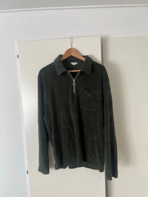 Mörkgrön långärmad tröja med dragkedja - Mörkgrön långärmad tröja i mjukt tyg med krage och halv dragkedja framtill. Tröjan har en bröstficka på vänster sida och avslappnad passform. Perfekt för lager-på-lager och chill dagar. Hör av er vid frågor!