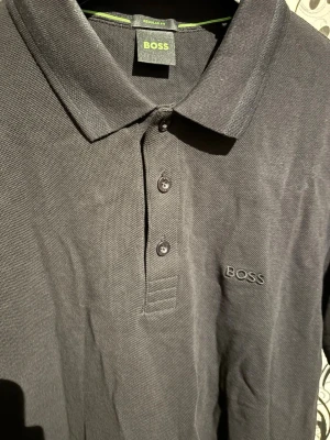 Svart Hugo Boss piketröja - Svart Hugo Boss piketröja i regular fit. Tillverkad i bomull med klassisk krage och knäppning framtill. Diskret broderad Hugo Boss-logotyp på bröstet. I mycket bra skick.