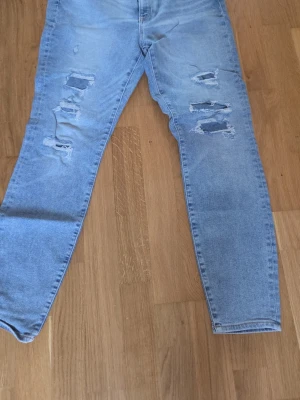 Ljusa super skinny jeans med slitningar - Säljer ett par ljusblå super skinny jeans med hög midja och ankellängd. Jeansen har flera slitningar och hål framtill för en trendig look. Tillverkade i denim och passar perfekt för dig som gillar en smal passform.